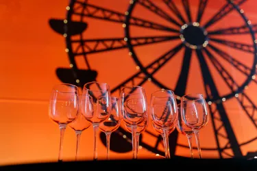 Eine Reihe von Weingläsern steht vor dem Hintergrund eines großen Riesenrades. Der Hintergrund ist in warmen Orange- und Schwarztönen gehalten.