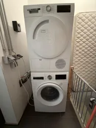 Ein kombiniertes Wasch- und Trockengerät in Weiß steht in einem Raum. Hinter dem Gerät ist eine Matratze und ein Geländer zu sehen.