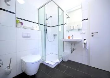 Ein modernes Badezimmer mit einer Duschkabine, einem Waschbecken und einer Toilette. Die Wände sind in schlichtem Weiß gefliest, und der Boden ist dunkel.