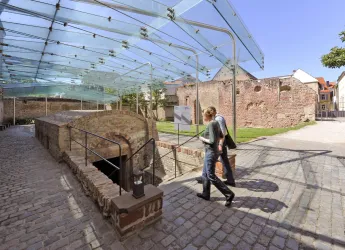 Ein archäologischer Ort mit Ruinen und einem modernen Glasdach. Zwei Personen spazieren auf einem gepflasterten Weg.