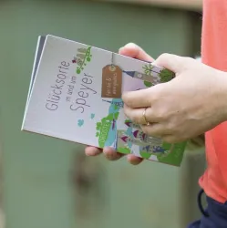 Eine Person hält ein Buch mit dem Titel "Glücksorte in und um Speyer". Das Buch ist bunt gestaltet und enthält Illustrationen von Sehenswürdigkeiten.