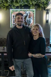 Ein Paar steht lächelnd in einem Restaurant mit einer gemütlichen Atmosphäre. Im Hintergrund ist ein dekoratives Schild zu sehen.