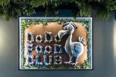 Ein kreatives Schild des "DoDo Food Club" zeigt einen Dodo-Vogel und den Schriftzug in reliefartigem Stil. Es ist von grünem Blattwerk umgeben und hat eine moderne Beleuchtung.