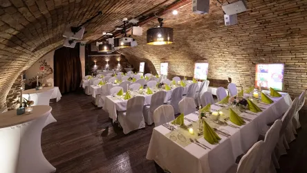 Ein eleganter Veranstaltungsraum mit Tischen, die in Weiß gedeckt sind. Die Wände sind aus Backstein und die Decke wird von stilvollen Lampen beleuchtet.
