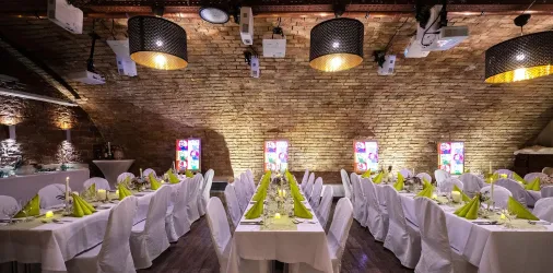 Ein eleganter Veranstaltungsraum mit langen Tischen, die mit weißen Tischdecken und grünen Servietten dekoriert sind. Die warmen Lichtakzente schaffen eine einladende Atmosphäre.