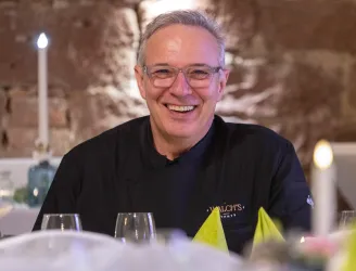 Ein lächelnder Koch in schwarzer Kleidung sitzt an einem schön gedeckten Tisch. Der Hintergrund ist mit warmem Licht und dekorativen Elementen gestaltet.