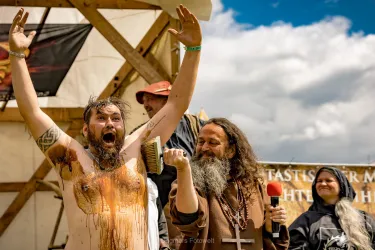 Ein Mann mit Bart und einem aufgeregten Gesicht feiert, während er mit einer Bürste über seinen Oberkörper gestrichen wird. Im Hintergrund stehen weitere Personen, die anscheinend an einer Veranstaltung teilnehmen.