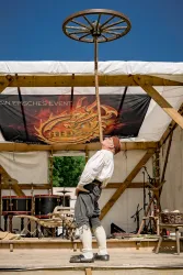 Ein Performer balanciert einen großen Wagenrad auf einem langen Stock. Die Bühne ist im Freien mit bunten Dekorationen und einer festlichen Atmosphäre.