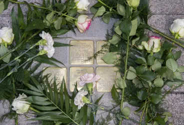 Eine Gedenkstätte mit mehreren Messingtafeln, umgeben von frischen Blumen. Die Blumen symbolisieren Erinnerung und Ehrfurcht.