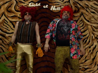 Zwei Männer in auffälligen, bunten Outfits liegen auf einem gestreiften Hintergrund. Sie tragen rote Perücken und Accessoires, die einen humorvollen Eindruck vermitteln.