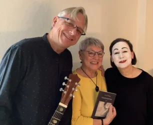 Drei Personen posieren gemeinsam für ein Foto. Eine Person hält ein Buch, während eine andere einen weißen Theatermakeup trägt.