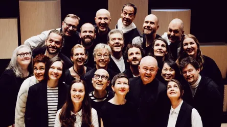 Eine Gruppe von Menschen steht in mehreren Reihen und lächelt in die Kamera. Sie tragen überwiegend schwarze und weiße Kleidung und befinden sich auf einer Bühne.