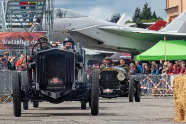 Zwei historische Autos fahren an einer Zuschauerreihe vorbei, während Menschen auf einer Veranstaltung zuschauen. Im Hintergrund ist ein Flugzeug und ein Zelt zu sehen.
