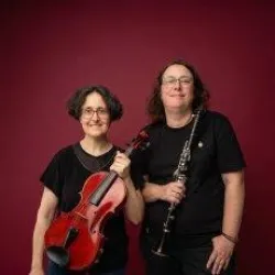 Zwei Musikerinnen stehen vor einem roten Hintergrund. Eine hält eine Geige, die andere eine Klarinette.