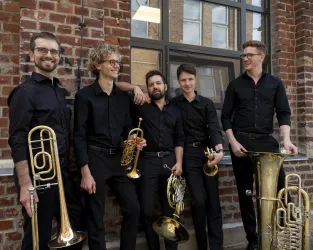 Eine Gruppe von fünf Musikern steht vor einer Backsteinwand. Sie tragen schwarze Kleidung und halten verschiedene Blasinstrumente.