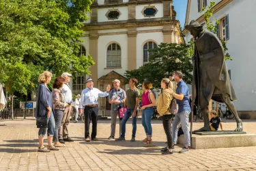 Eine Gruppe von Touristen hört einem Reiseführer zu, während sie auf einem Platz stehen. Im Hintergrund ist eine Statue und historische Gebäude zu sehen.