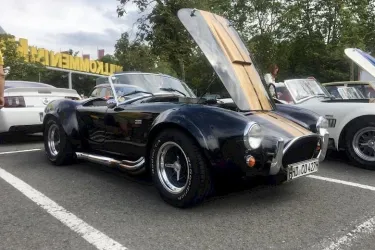 Ein schwarzer Sportwagen mit offenem Motorhaube steht auf einem Parkplatz. Im Hintergrund sind weitere Autos und Bäume zu sehen.