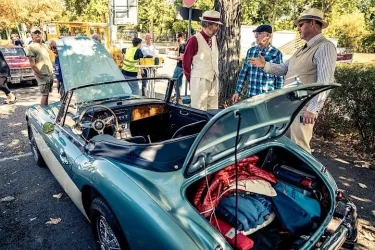 Eine Gruppe von Männern in stilvollen Outfits steht um ein klassisches Auto. Der Kofferraum des Fahrzeugs ist geöffnet und zeigt Gepäck.