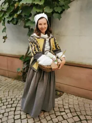 Eine lächelnde Frau in traditioneller Kleidung hält einen Korb. Sie steht auf einem gepflasterten Weg neben einer Mauer mit Pflanzen.