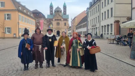 Sechs Personen in historischer Kleidung stehen auf einem Platz. Im Hintergrund sind alte Gebäude und ein Kirchturm zu sehen.