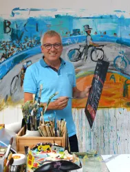 Ein lächelnder Mann in einem blauen Poloshirt steht in einem künstlerischen Atelier. Im Hintergrund ist ein farbenfrohes Gemälde mit Fahrrädern sichtbar, während er einen Pinsel in der Hand hält.