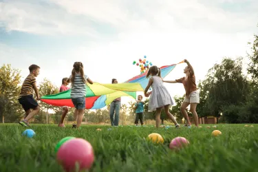 Eine Gruppe von Kindern spielt im Freien mit einem bunten Tuch. Bunte Bälle liegen auf dem grünen Gras.