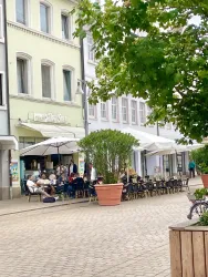 Eine belebte Straßencafé mit Tischen und Stühlen unter großen Sonnenschirmen. Menschen sitzen draußen und genießen die Atmosphäre.