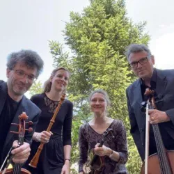 Eine Gruppe von vier Musikern steht im Freien vor einem großen Baum. Sie halten ihre Instrumente und lächeln in die Kamera.