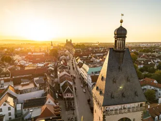 Eine malerische Stadtansicht bei Sonnenuntergang, mit einem Blick auf historische Gebäude und Straßen. Der Himmel leuchtet in warmen Farben über den Dächern.