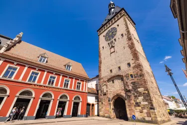 Ein historischer Turm mit einer Uhr in einer Stadt. Im Vordergrund sind schöne Gebäude und ein strahlend blauer Himmel zu sehen.