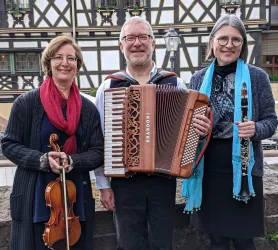 Drei Musiker stehen lächelnd zusammen. Sie halten verschiedene Musikinstrumente: eine Violine, ein Akkordeon und ein Klarinette.