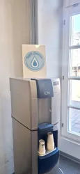 Ein Wasserspender steht in einem hellen Raum neben einem Fenster. Oben auf dem Spender befindet sich ein Schild mit einem Wassertropfen-Logo.