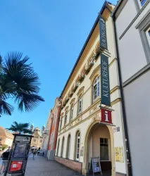 Ein historisches Gebäude mit einer Werbetafel für das Kulturhaus. Im Vordergrund sind einige Palmen und eine entspannte Stadtatmosphäre zu sehen.