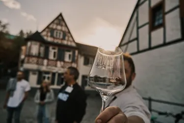 Ein Mann hält ein Weinglas vor einem historischen Gebäude. Im Hintergrund sind weitere Personen zu sehen, während die Sonne untergeht.