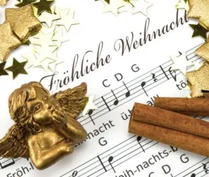 Ein goldener Engel sitzt auf Noten mit Weihnachtsliedern. Daneben sind Zimtstangen und goldene Sterne verstreut.