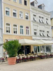 Ein einladendes Café mit Außenbereich und einem großen Topf mit Pflanzen. Die Fassade ist hell und die Fenster sind teils geöffnet.