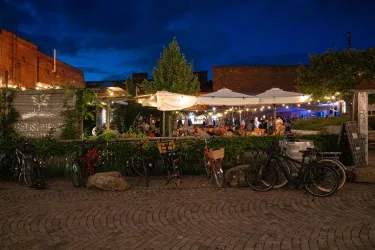 Ein belebter Außenbereich eines Restaurants bei Nacht, umgeben von Bäumen und Pflanzen. Fahrradständer sind im Vordergrund sichtbar, während Gäste im Hintergrund entspannen.