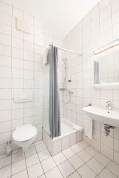 Ein modernes Badezimmer mit weißen Fliesen und einer Dusche. Es gibt ein Waschbecken und eine Toilette, die sauber und ordentlich aussehen.