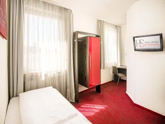 Ein einfaches Hotelzimmer mit einem Einzelbett, einem Kleiderschrank und einem Schreibtisch. Große Fenster sorgen für viel Tageslicht.
