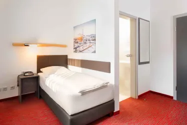 Ein gemütliches Hotelzimmer mit einem Einzelbett und einem modernen Design. Der Boden ist rot und es gibt ein Bild an der Wand sowie Zugang zu einem Badezimmer.