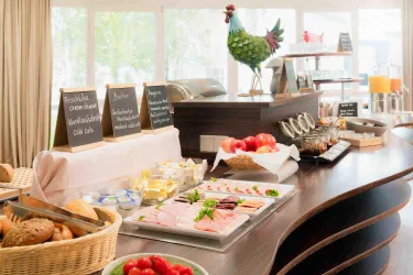 Ein Frühstücksbuffet mit verschiedenen Lebensmitteln wie Brot, Wurst, Käse und frischem Obst. Im Hintergrund steht eine dekorative Henne auf dem Tisch.