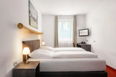 Ein modernes Schlafzimmer mit einem Doppelbett und zwei Nachttischlampen. Durch das Fenster fällt Tageslicht und die Wände sind hell gestaltet.