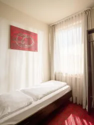Ein einfaches Schlafzimmer mit einem Einzelbett und einem roten Teppich. Das Fenster ist mit hellen Vorhängen dekoriert und lässt viel Tageslicht herein.