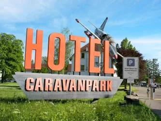 Ein Schild mit der Aufschrift "Hotel Caravanpark" und einem Flugzeugmodell darüber. Die Umgebung ist grün und sonnig.