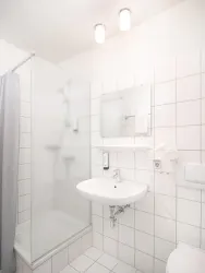 Ein modernes Badezimmer mit einer Dusche, Waschbecken und einem großen Spiegel. Die Wände sind mit weißen Fliesen verkleidet und der Raum ist hell beleuchtet.