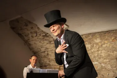 Ein Mann trägt einen Zylinder und einen Anzug, während er auf einer Bühne performt. Im Hintergrund sitzt ein Pianist und spielt Musik.