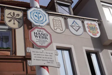 Eine Reihe von Wappen und Schildern hängt an einer Wand. Das zentrale Schild weist auf das Handwerk hin und enthält Informationen über die Bedeutung dieses Gewerbes.