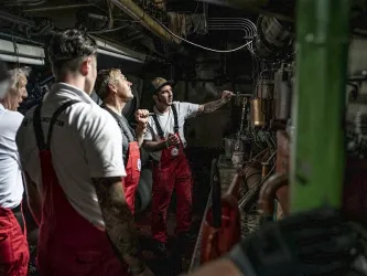 Eine Gruppe von Arbeitern in roter Arbeitskleidung steht in einem Maschinenraum. Sie schauen auf eine Maschine und scheinen ein technisches Problem zu besprechen.
