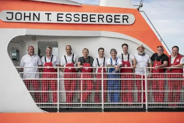 Eine Gruppe von Männern in roten Overalls steht auf einem Schiff mit der Aufschrift "JOHN T. ESSBERGER". Sie lächeln und posieren gemeinsam auf dem Vorderdeck.
