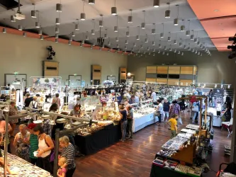 Ein belebter Markt in einem großen Raum mit vielen Ständen. Menschen schlendern umher und schauen sich verschiedene Waren an.